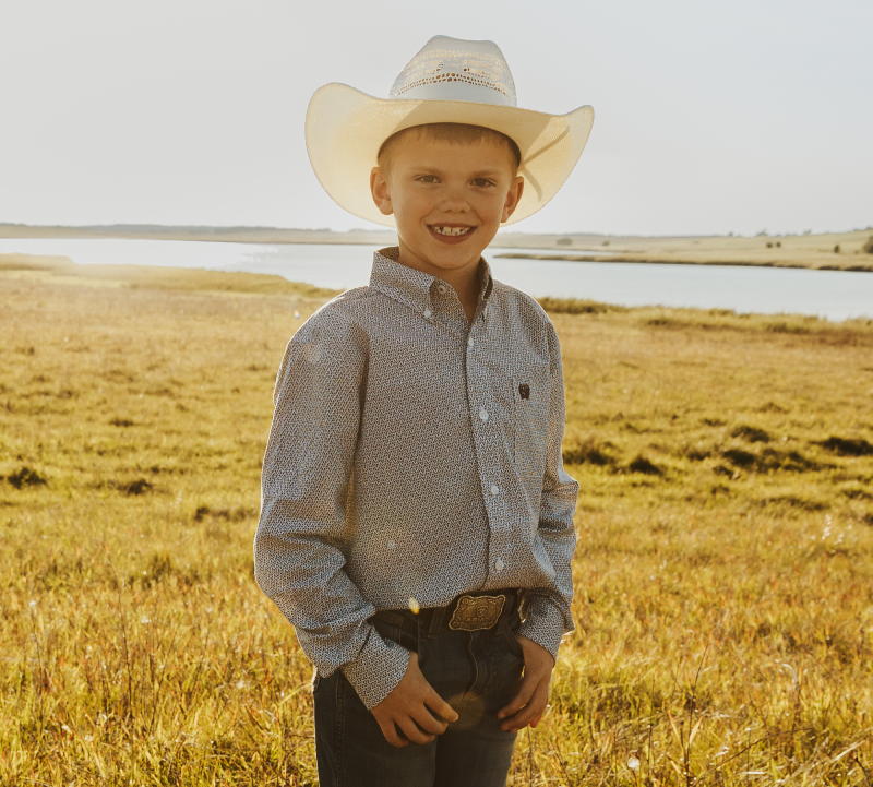 Young boy with black best and long sleeve white shirt standing to the right of woman with shoulder length brown hair and short sleeve white blouse in a pasture with a pond in the background