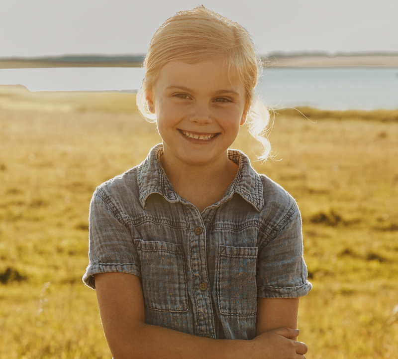 Young girl with shoulder length blonde hair and white bow in a light blue short sleeve blouse standing in a pasture with a pond and blue sky in the background