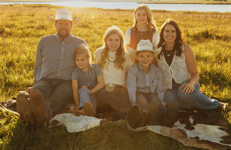 Black and white photo of a dad kneeling in a pasture with two of his daughters standing on each side of him and youngest daughter sitting on his knee
