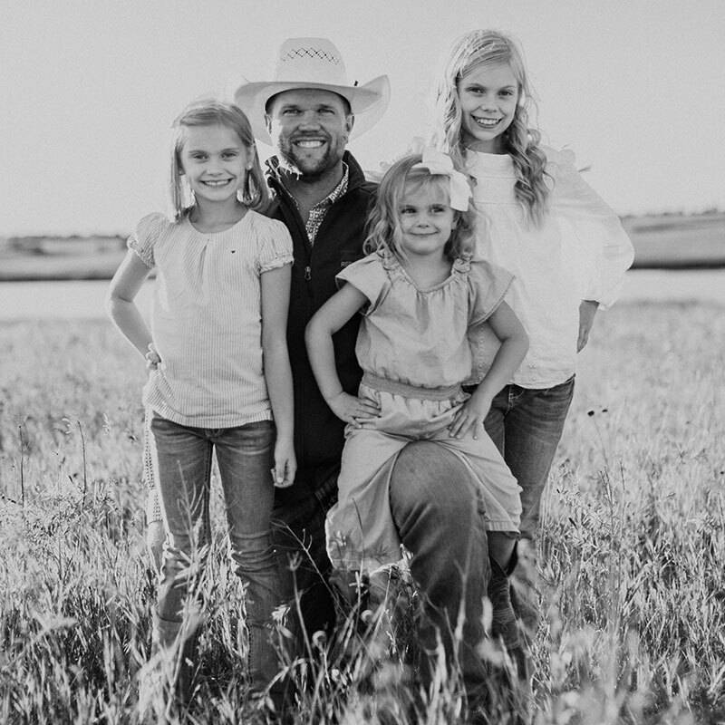 Black and white photo of a dad kneeling in a pasture with two of his daughters standing on each side of him and youngest daughter sitting on his knee